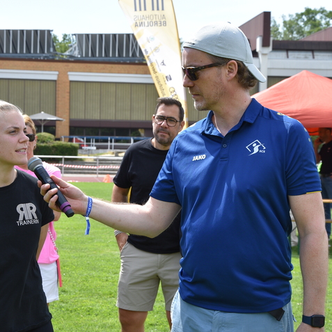Interview mit einer Trainerin im Sportfeld, w&auml;hrend ein Moderator mit Mikrofon spricht. Zuschauer im Hintergrund.