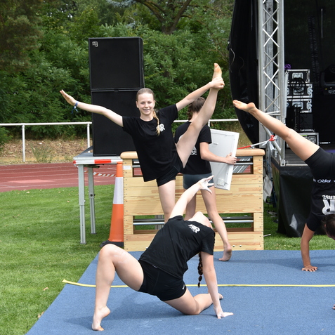 Mehrere Turnerinnen in schwarzen Shirts f&uuml;hren eine Akrobatik&uuml;bung auf blauer Matte im Freien vor.