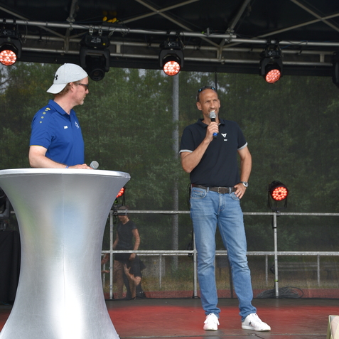Zwei M&auml;nner stehen auf einer B&uuml;hne; einer spricht ins Mikrofon, der andere beobachtet, im Hintergrund sind Lichter sichtbar.
