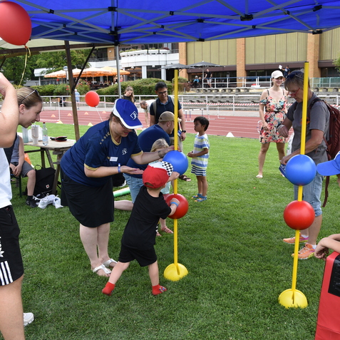 Kinder spielen mit farbigen B&auml;llen und einer Betreuerin unter einem Zelt auf einer Wiese bei einer Veranstaltung.