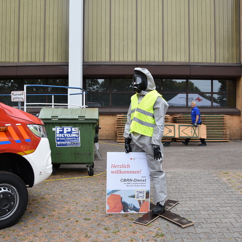 Sicherheitsmannikins in Schutzanzug steht vor einem Gebäude, während ein Mitarbeiter Tische trägt. Rettungsfahrzeug im Vordergrund.