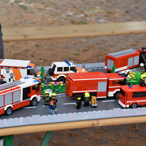 Detailreiche LEGO-Szene mit Feuerwehrfahrzeugen, Rettungsdienst und Minifiguren auf einer Straße mit Blumen.