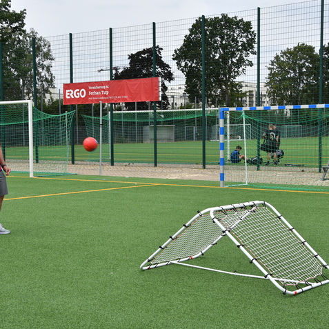 Mann mit buntem Trikot steht auf einem Kunstrasenplatz und wirft einen roten Ball auf ein Sportnetz.