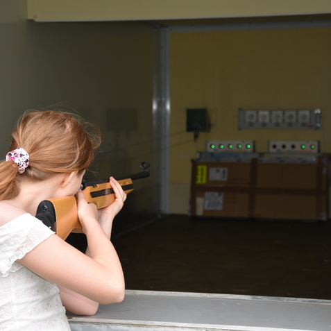 Mädchen mit einem Gewehr zielt auf eine Zielscheibe in einem Schießstand. Hinter der Zielscheibe sind Kontrollleuchten sichtbar.