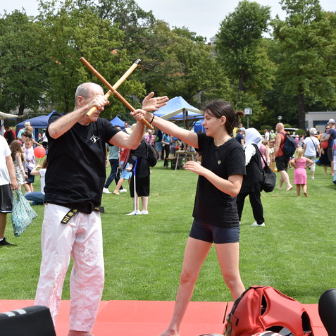 Zwei Personen trainieren mit Bambus-Stöcken in einem Park, umgeben von Zuschauern und Marktständen.