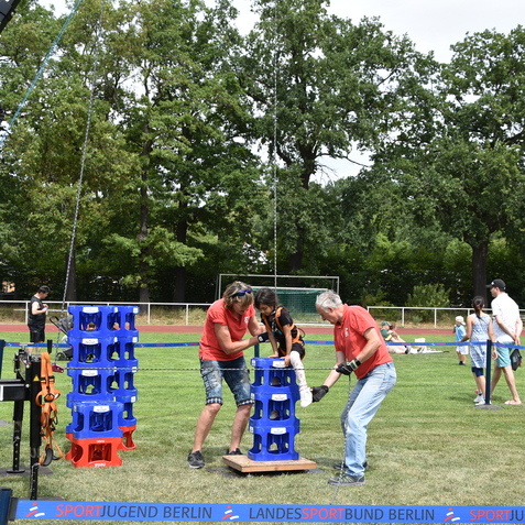 Zwei Erwachsene unterstützen ein Kind beim Klettern auf einen blauen Kastenstapel auf einem Sportplatz.