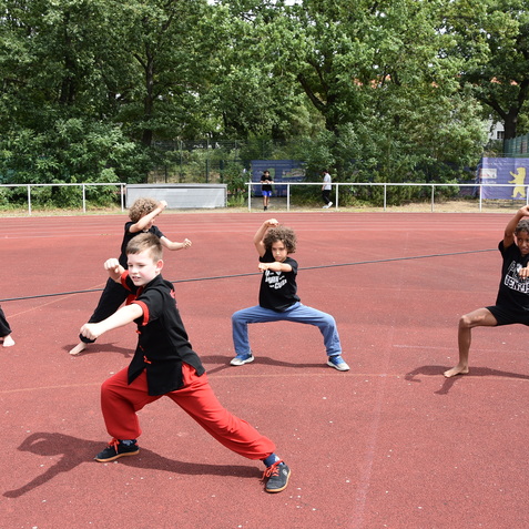Gruppe von Kindern in schwarzen T-Shirts, die auf einem roten Sportplatz Kampfbewegungen ausführen.