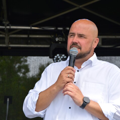 Mann mit Glatze spricht in ein Mikrofon, trägt ein weißes Hemd und steht auf einer Bühne mit grünem Hintergrund.