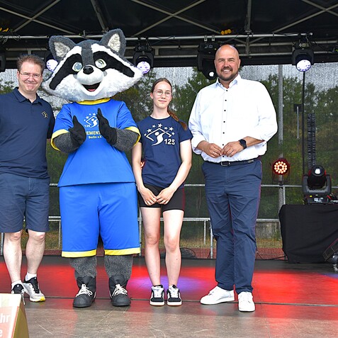 Drei Personen und ein Maskottchen stehen auf einer B&uuml;hne, umgeben von Lichtern und einem Tontechnik-Setup.