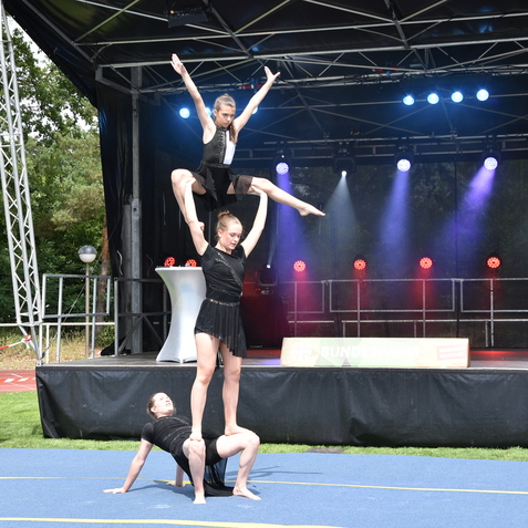 Drei Artistinnen in Schwarz zeigen eine akrobatische Pyramide auf einer blauen Fl&auml;che vor einer B&uuml;hne mit Beleuchtung.
