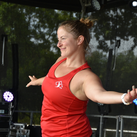 Frau in rotem Sporttops tanzt auf einer B&uuml;hne, hinter ihr unscharfe Lichter und schwarze Vorh&auml;nge.