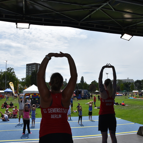 Zwei Trainer in roten Shirts demonstrieren eine &Uuml;bung vor einer Gruppe von Kindern auf einer Sportfl&auml;che im Freien.