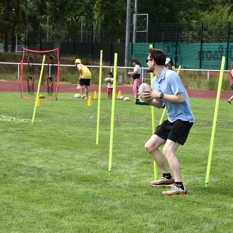Sportliche &Uuml;bung auf einem Grasplatz, mehrere Kinder und Erwachsene halten Rugbyb&auml;lle und trainieren mit H&uuml;tchen.