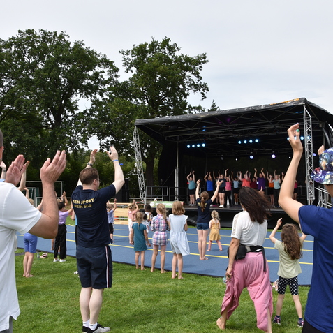 Gruppe von Menschen auf einer Wiese, die zu einer Tanzauff&uuml;hrung auf einer B&uuml;hne mit bunten Lichtern mitklatschen.