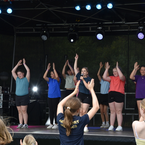 Gruppe von Kindern auf einer B&uuml;hne, die mit erhobenen H&auml;nden tanzen und eine S&auml;ngerin mit Mikrofon in der Mitte.