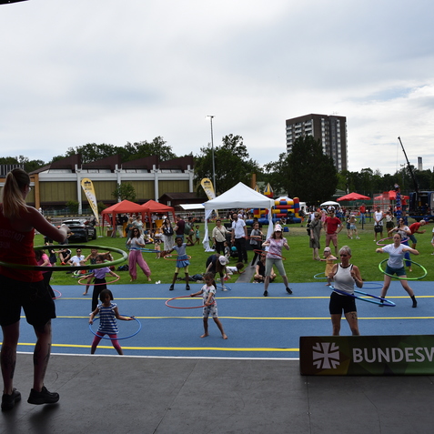 Gruppen von Kindern und Erwachsenen tanzen mit Hula-Hoops auf einem bunten Sportplatz bei einer Veranstaltung.