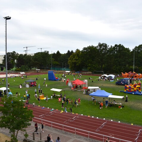 Blick auf ein Festgel&auml;nde mit Menschen, St&auml;nden, einer H&uuml;pfburg und einer Laufbahn umgeben von B&auml;umen.