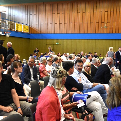 Vielbesch&auml;ftigte Zuschauer sitzen in einem Sporthallenraum, w&auml;hrend sie miteinander sprechen und Unterlagen in H&auml;nden halten.