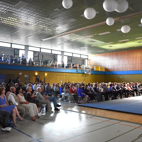 Eine Vielzahl von Menschen sitzt auf St&uuml;hlen in einer Sporthalle w&auml;hrend einer Veranstaltung.