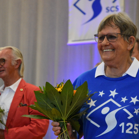 L&auml;chelnde Frau in blauer T-Shirt mit dem Schriftzug &bdquo;125&ldquo; h&auml;lt einen Strau&szlig; Blumen, umgeben von weiteren Personen auf einer Feier.