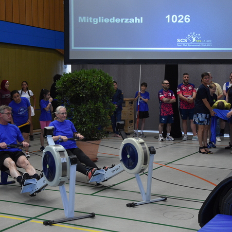 Zwei Personen beim Rudern auf Ergometern in einer Sporthalle, umgeben von Zuschauern in blauen T-Shirts.