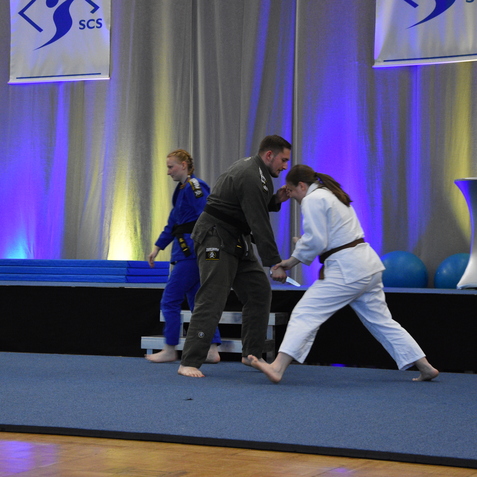 Zwei Judoka in Wettkampfbekleidung trainieren auf einer blauen Matte, w&auml;hrend Zuschauer im Hintergrund beobachten.