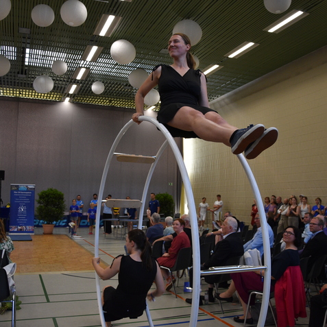 Frau sitzt auf einem gro&szlig;en Reckring, w&auml;hrend das Publikum in einem Sportzentrum zuschaut. Stimmungsvolle Beleuchtung im Hintergrund.