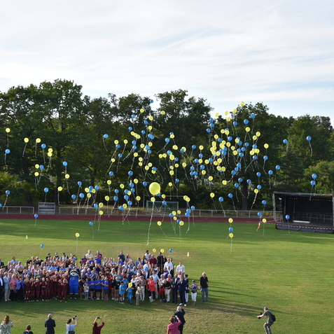 Gruppe von Menschen auf einem Sportplatz, w&auml;hrend bunt gef&uuml;llte Ballons in den Himmel steigen.