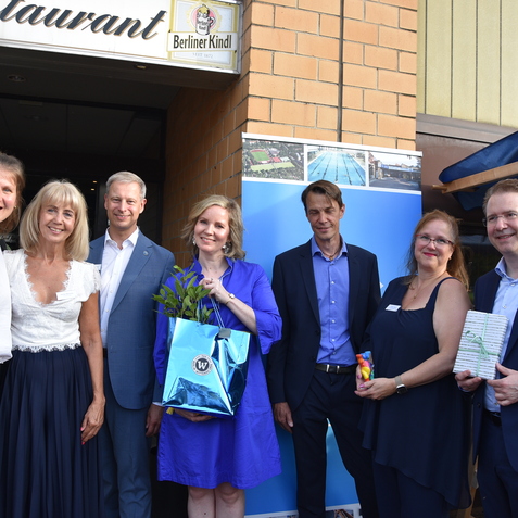 Gruppe von sieben Personen vor einem Restaurant mit blauen Bannern, eine Person h&auml;lt einen Geschenkkorb mit Pflanzen.