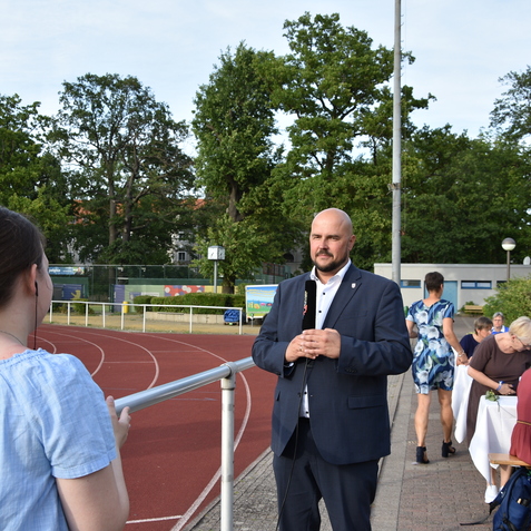 Mann im Anzug spricht auf einem Sportplatz, w&auml;hrend eine Frau ihm interessiert zuh&ouml;rt. Im Hintergrund eine Tischreihe mit G&auml;sten.