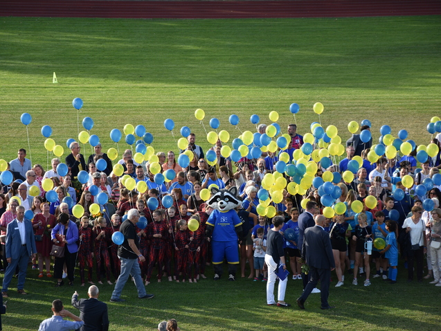 Gruppenszene auf einem Rasen mit Menschen, die gelbe und blaue Luftballons halten, während ein Maskottchen in der Mitte steht.