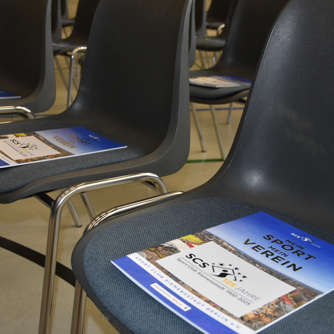 Reihe von Stühlen mit Prospekten des Sportvereins auf den Sitzflächen in einem Innenraum.
