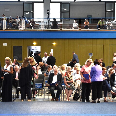 Menschen bei einer Veranstaltung in einer Halle, einige stehen, andere sitzen auf Sesseln und unterhalten sich.