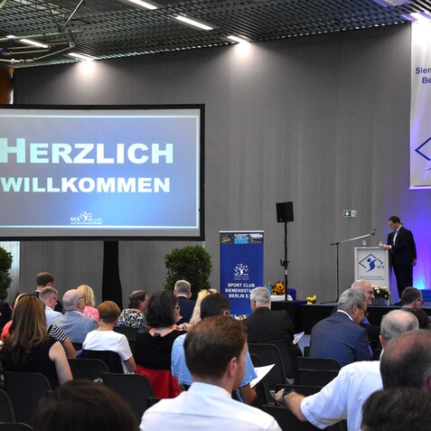 Veranstaltungshalle mit Publikum und Pr&auml;sentation &bdquo;Herzlich Willkommen&ldquo; auf gro&szlig;er Leinwand, im Hintergrund ein Redner.