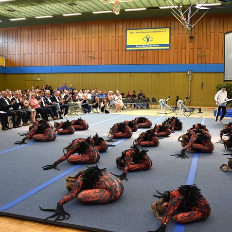 Tanzgruppe in roten Kost&uuml;men mit Federkopfschmuck auf einer B&uuml;hne, Publikum applaudiert im Hintergrund.
