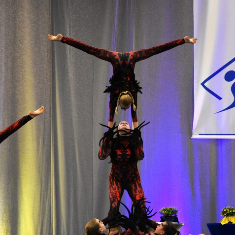 Akrobatische Aufführung: Eine Sportlerin steht auf einem anderen und balanciert eine dritte Person mit erhobenen Händen.