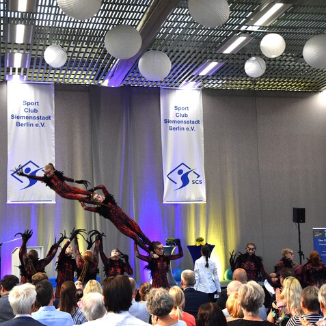 Tanzauff&uuml;hrung in einem Sportclub, mit Akteuren in bunten Kost&uuml;men und einer anfeuernden Menge im Hintergrund.