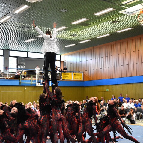K&uuml;nstlerische Darbietung: T&auml;nzer bilden eine Pyramide, w&auml;hrend Zuschauer in einem Sporthalle zuschauen.
