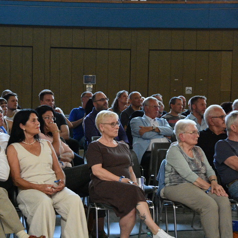 Zahlreiches Publikum sitzt auf St&uuml;hlen in einer Veranstaltungsraum, fasziniert und aufmerksam.