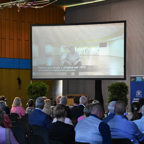 Bild einer Veranstaltung mit Publikum, w&auml;hrend ein Video &uuml;ber Sabine zum Bruch auf einer Leinwand gezeigt wird.