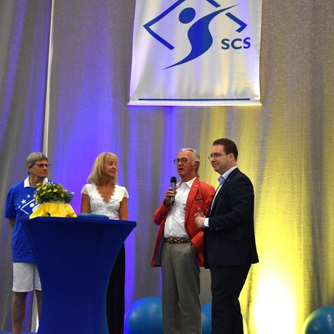 F&uuml;nf Personen stehen an Stehtischen auf einer Veranstaltung, ein Redner h&auml;lt ein Mikrofon, B&uuml;hnenbanner im Hintergrund.