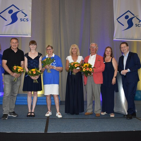 Sieben Personen stehen auf einer B&uuml;hne, halten Blumenstr&auml;u&szlig;e und l&auml;cheln, w&auml;hrend ein Banner mit dem Logo der SCS im Hintergrund h&auml;ngt.