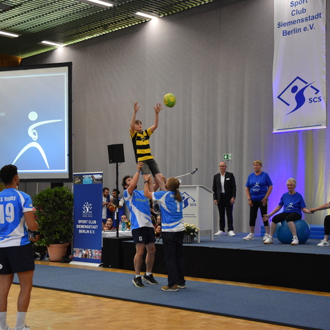 Kind in gelb-schwarz gestreiftem Shirt wird von Sportlern in blauen Trikots im Rugby hochgehoben. Im Hintergrund ist eine Pr&auml;sentation.