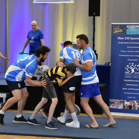 Gruppe von M&auml;nnern mit Rugbyball um einen Jungen, w&auml;hrend Frauen im Hintergrund auf einer Trainingsmatte sitzen.