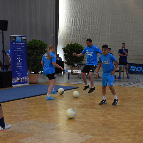 Vier Personen in blauen T-Shirts jonglieren mit Fu&szlig;b&auml;llen in einer Sporthalle. Im Hintergrund steht eine Informationswand.