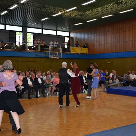 Tanzende Paare auf einer Veranstaltung in einer Sporthalle, Zuschauer sitzen in Reihen im Hintergrund.