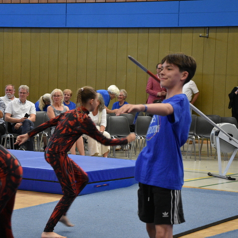 Junge in blauer Sportkleidung jongliert mit einem langen Stock, w&auml;hrend andere Gymnastinnen auf einer Matte trainieren.