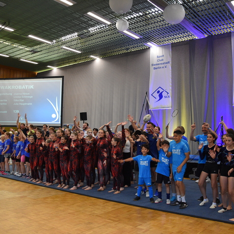 Gruppenszene mit T&auml;nzern in Kost&uuml;men, die auf einer B&uuml;hne in einem Sportzentrum jubeln und applaudieren.