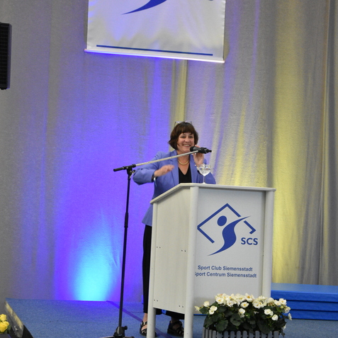 Frau am Rednerpult mit Mikrofon im Sportzentrum Siemensstadt, bl&uuml;hende Pflanzen im Vordergrund und Beleuchtung im Hintergrund.