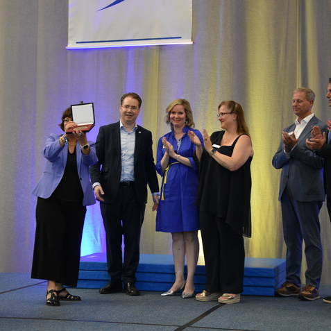 Eine Person h&auml;lt eine Auszeichnung hoch, w&auml;hrend eine Gruppe von Personen applaudiert und l&auml;chelt. B&uuml;hne mit SCS-Logo im Hintergrund.